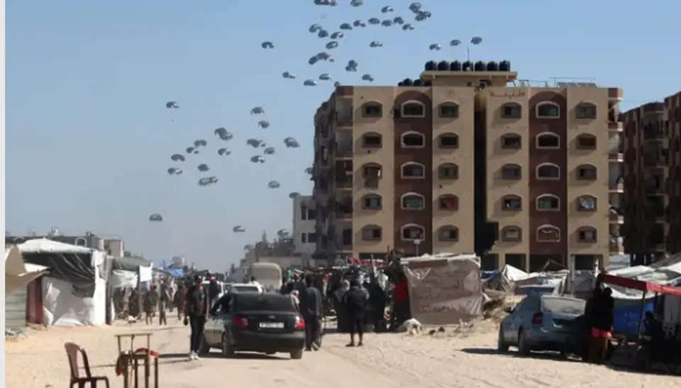 Aid parcels parachuting over a crowded street with tents and buildings in Gaza